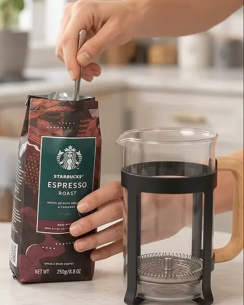 Cuchara grande tomando café molido de la bolsa junto a la prensa vacía.